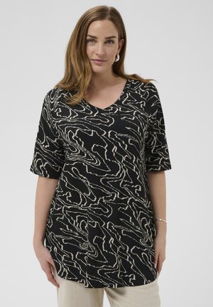 Femme aux cheveux châtain clair portant une tunique noire avec des motifs abstraits en lignes beige et un pantalon beige, debout devant un fond gris uni.