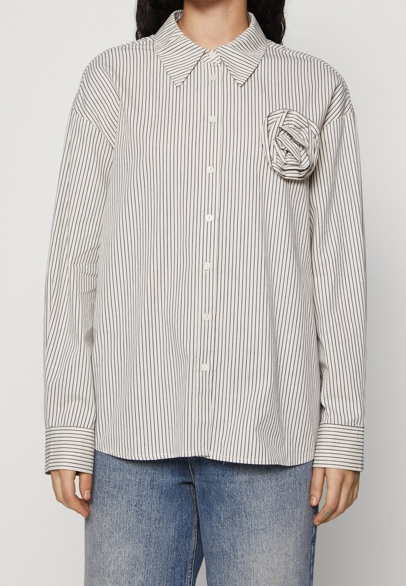Femme portant une chemise blanche boutonnée avec de fines rayures verticales noires et une rose en tissu assortie sur la poitrine gauche, associée à un jean bleu.