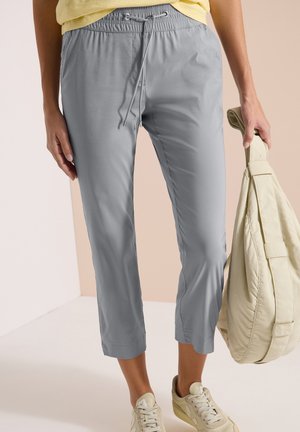 Persoon draagt lichtgrijze cropped broek met trekkoord, beige sneakers, gele top, houdt grote beige tas vast in een neutrale binnenomgeving.