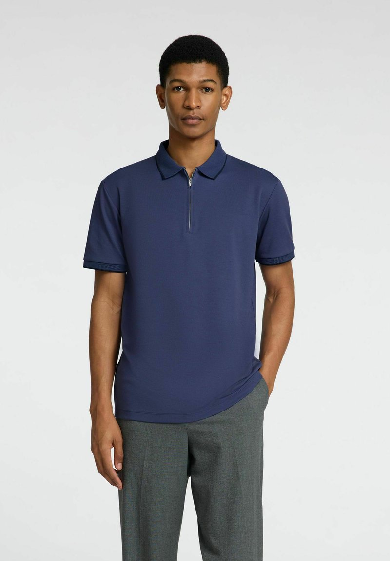 Polo marine à demi-fermeture éclair, manches courtes et col texturé. Porté avec un pantalon gris, présentant une coupe lisse et moderne.