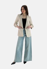 Blazer beige con vestibilità ampia e maniche arrotolate, abbinato a jeans a gamba larga blu chiaro e scarpe nere con punta affusolata. Top nero sotto.