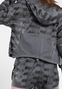 Femme portant un ensemble coordonné en gris composé d'une veste et d'un short Nike Air avec des motifs de logo noirs, montrant le dos avec une poche zippée sur la veste.