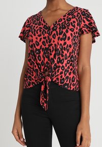 Blusa de estampado de leopardo rojo con mangas cortas y acampanadas, escote en V, botones frontales y detalle de nudo atado en la cintura, combinada con pantalones negros.