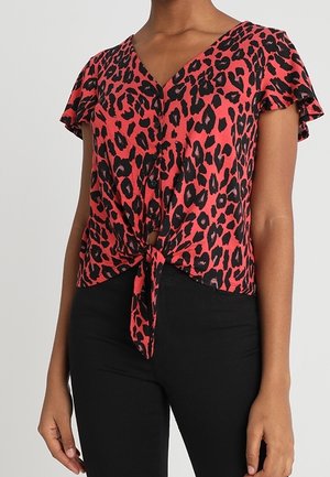 Blusa rossa con stampa leopardata, maniche corte a volant, scollatura a V, bottoni frontali e dettaglio a nodo in vita, abbinata a pantaloni neri.