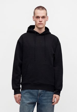Jeune homme aux cheveux courts portant un sweat à capuche noir avec poche sur le devant et jean bleu, debout devant un fond uni.