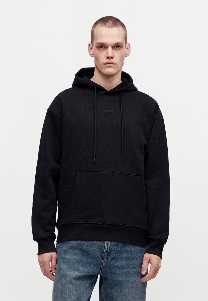Jeune homme aux cheveux courts portant un sweat à capuche noir avec poche sur le devant et jean bleu, debout devant un fond uni.