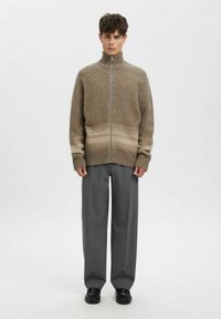 Bēšas krāsas adīts rāvējslēdzēja džemperis ar ribotu aproču un apakšmalu, ar smalku gradienta dizainu. Kombinēts ar pelēkām biksēm un melniem apaviem.