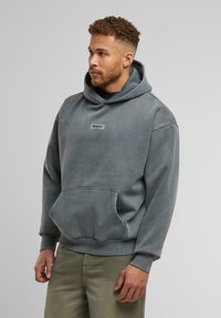 Grau Hoodie mit einer Fronttasche und gerippten Bündchen. Mit einem kleinen Etikett auf der Brust und einer großen Kordelzugkapuze. Weiche Textur.