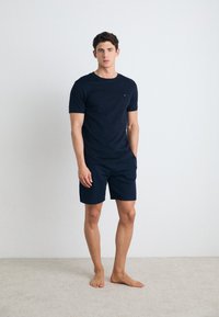 Camiseta de algodón azul marino de manga corta con un pequeño logo en el pecho, combinada con pantalones cortos a juego en azul marino, de pie sobre una alfombra clara.