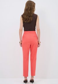 Sleeveless brown top paired with coral tapered trousers, featuring a smooth texture and a back pocket detail. High-heeled shoes complete the look.
