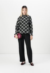 Vrouw in een blouse met zwart-wit diamantpatroon, zwarte pants, bordeauxrode flats en een bordeauxrode clutch tegen een effen achtergrond.