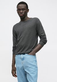 Νεαρός άνδρας με σκούρο δέρμα, φορώντας ένα σκούρο γκρι πουλόβερ με μακριά μανίκια και ανοιχτό μπλε τζιν, στέκεται με το ένα χέρι στην τσέπη μπροστά σε ένα απλό φόντο.