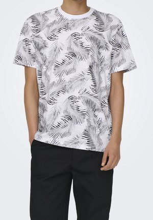 Mężczyzna w białej koszulce z krótkim rękawem z czarnym i szarym nadrukiem liści palmy oraz czarnych spodniach, stoi z jedną ręką w kieszeni.