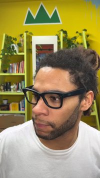 Lunettes rectangulaires noires à monture épaisse, portées par une personne aux cheveux foncés et bouclés. En arrière-plan, des murs jaunes éclatants et de la verdure.