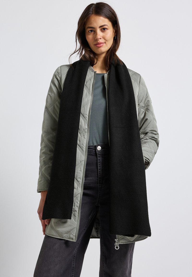 Giacca verde oliva trapuntata con chiusura a zip e sciarpa in lana nera a coste. Parte superiore grigia e pantaloni di denim scuro completano il look.