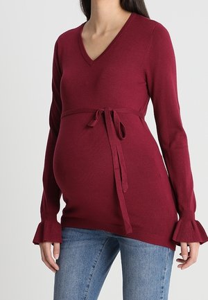 Gravid kvinde iført en langærmet, burgunder rød V-hals sweater med bindebånd i taljen og flæser ved manchetterne, kombineret med blå jeans.