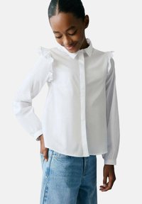 Cămașă albă cu mâneci lungi, cu detalii cu volane pe umeri, având un guler clasic și închidere cu nasturi, asortată cu blugi blue.