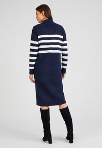 Abito a maglia blu navy con strisce orizzontali bianche, presenta un collo a camicia, maniche lunghe e una lunghezza fino al polpaccio. Texture morbida.