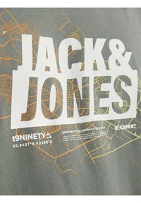 Camiseta gris con el texto en blanco "JACK & JONES" y un diseño de mapa abstracto en naranja-amarillo, con detalles de construcción impresos abajo.