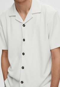 Man wearing a short-sleeve white ribbed shirt with dark buttons and a camp collar, hands partially in pockets, cropped at chest and waist.