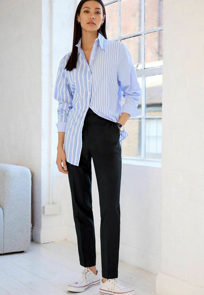 Stribet blå og hvid skjorte med knapånd, i en afslappet pasform, parret med sorte tapered bukser og hvide sneakers. Glat tekstur, casual stil.