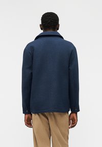 Μπλε ναυτικό σακάκι από μείγμα μαλλιού με γιακά, μακριά μανίκια και μανσέτες με κουμπιά, εμφανιζόμενο από πίσω σε ανοιχτό φόντο.