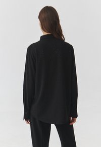 Maglietta nera a maniche lunghe con un design texturizzato, caratterizzata da accenti sparkling e un colletto tradizionale. Tessuto morbido, vestibilità rilassata e polsini con bottone.