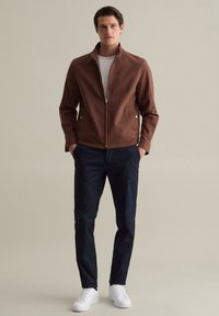 Braune Zip-Jacke mit hohem Kragen, getragen über einem weißen Hemd, kombiniert mit marineblauen Hosen und weißen Sneakers. Glatte Textur, lässiges Design.