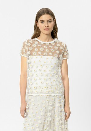 Junge Frau trägt ein weißes, durchsichtiges Oberteil und einen Rock, die mit 3D-Margaritenblumen-Applikationen verziert sind, und steht vor einem schlichten weißen Hintergrund.