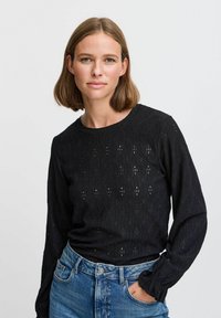 Haut noir à manches longues avec un motif texturé comportant des découpes en losange, un col rond et des poignets élastiqués. Associé à un jean en denim bleu.