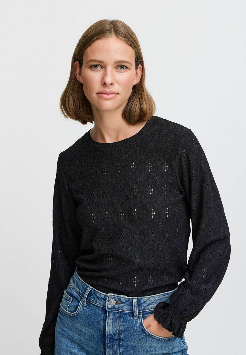 Haut noir à manches longues avec un motif texturé comportant des découpes en losange, un col rond et des poignets élastiqués. Associé à un jean en denim bleu.