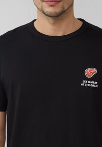 Czarny bawełniany t-shirt z okrągłym dekoltem, krótki rękaw i białym haftem przedstawiającym grafikę mięsa oraz tekst "SPOTKAJMY SIĘ NA GRILLU!"
