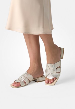 Pieds chaussés de sandales plates en cuir tressé argenté métallique, à talons blocs bas et bouts carrés, assorties à une jupe beige clair.