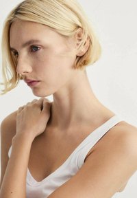 Hvid tanktop på en model med kort blond hår, neutral hudtone og subtile fregner, der poser med en hånd på sin skulder.