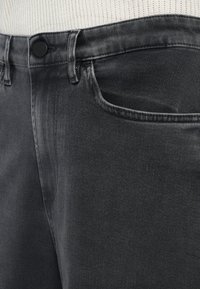 Közeli felvétel fekete farmernadrág zsebének és derékpántjának fekete gombbal, amely egy fehér bordás kötött felső fölött van viselve.