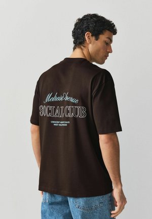 Junger Mann mit lockigem dunklem Haar trägt ein schwarzes T-Shirt mit der Aufschrift „Melrose Avenue Social Club, Crescent Heights Blvd, West Hollywood“ kombiniert mit blauen Jeans.