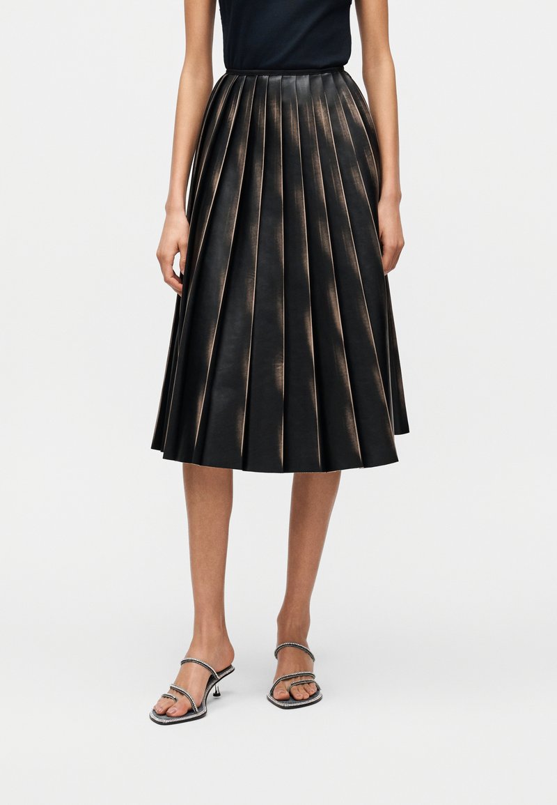 Μαύρη πλισέ φούστα από δέρμα, με κάθετες πιέτες που δημιουργούν υφή. Συνδυάζεται με μαύρα σανδάλια με λουράκια και διακοσμητικές λεπτομέρειες.