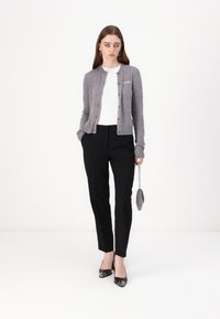 Grå strikket cardigan med knapper, hvit skjorte, svarte skreddersydde bukser og svarte lakkhæler, som holder en liten metallisk veske.