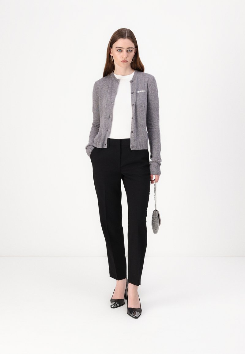 Graue Strickjacke mit Knöpfen, weißes Hemd, schwarze maßgeschneiderte Hose und schwarze Lack-Pumps, hält eine kleine metallische Tasche.
