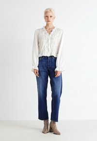 Blusa branca com pregas e mangas longas, combinada com jeans soltos azuis e botins bege. Textura suave, design casual.