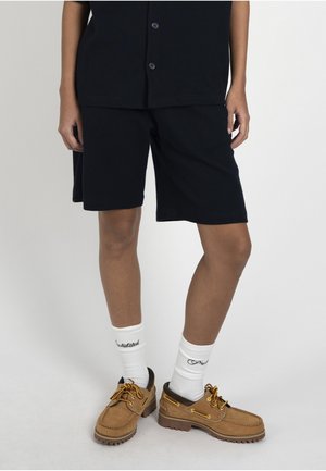 Osoba ubrana w czarne spodenki i koszulkę, białe skarpetki za kostkę z napisem oraz jasnobrązowe wiązane buty żeglarskie na jasnym tle.