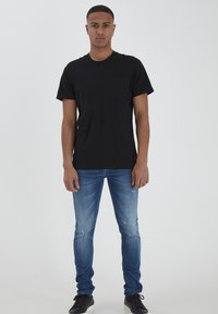 Juoda medvilninė t-shirt su trumpomis rankovėmis ir krūtine kišene, derinama su nublukusiais mėlynais džinsais ir juodais sportbačiais, prieš baltą foną.