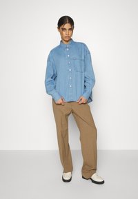 Camicia di jeans blu chiaro con design a bottoni, taschino sul petto e vestibilità comoda, abbinata a pantaloni dritti color beige e scarpe bianche.