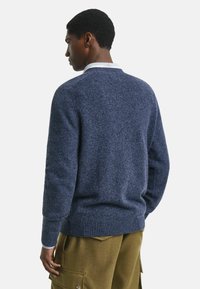 Junger Mann, von hinten gesehen, trägt einen blauen Pullover über einem weißen Kragenhemd und olivfarbene Cargohosen vor einem einfarbigen Hintergrund.