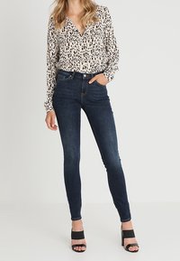 Mujer con jeans skinny de color azul oscuro, blusa beige con estampado de leopardo y sandalias de tacón negro, de pie con una mano en el bolsillo.