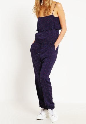 Vrouw draagt een marineblauwe mouwloze jumpsuit met kleine rode patronen en witte sneakers, staand met handen in de zakken.