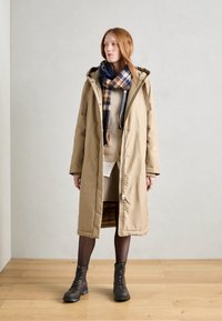 Abrigo acolchado largo y beige con capucha, cierre de cremallera y botones a presión. Lleva un suéter crema debajo, combinado con medias negras y botas con cordones. Accesorio con una bufanda a cuadros en azul y naranja.