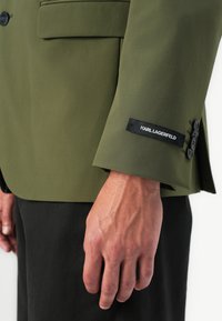 Close-up of a person’s hand beside the sleeve of an olive green blazer with black buttons and a Karl Lagerfeld label.