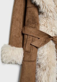 Brun ruskindsfrakke med en kunstig pelskrave og manchetter. Har et stofbælte og en blød tekstur, der giver et varmt og hyggeligt udseende.