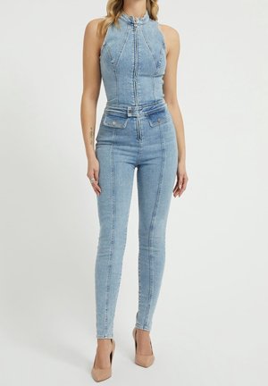 Kvinde iført lyseblåt denim ærmeløst jumpsuit med lynlås foran og bælte, kombineret med beige højhælede sko, står foran en hvid baggrund.